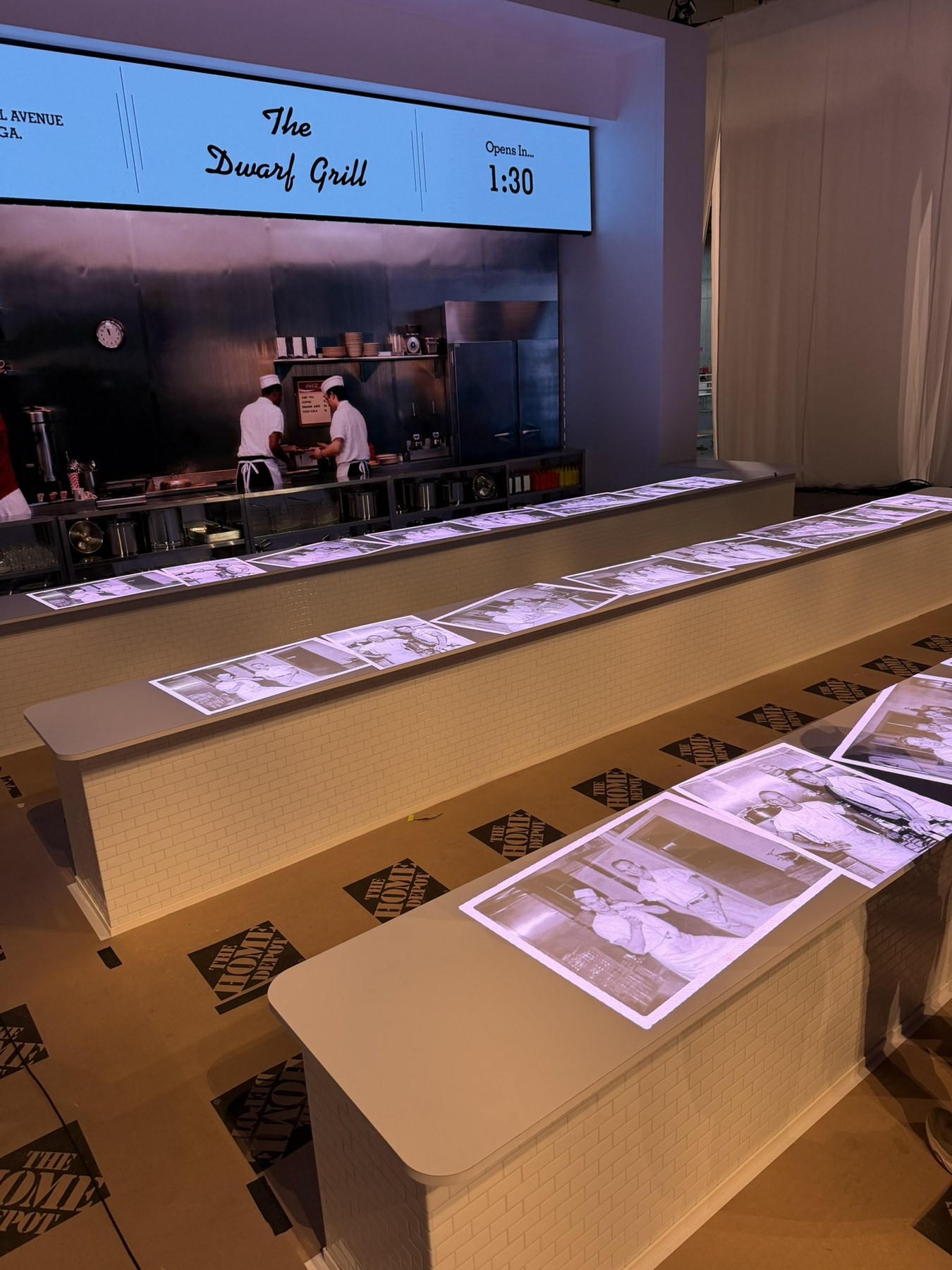 Archival photographs projected along three white dining counters, with a film playing on the screen behind them.