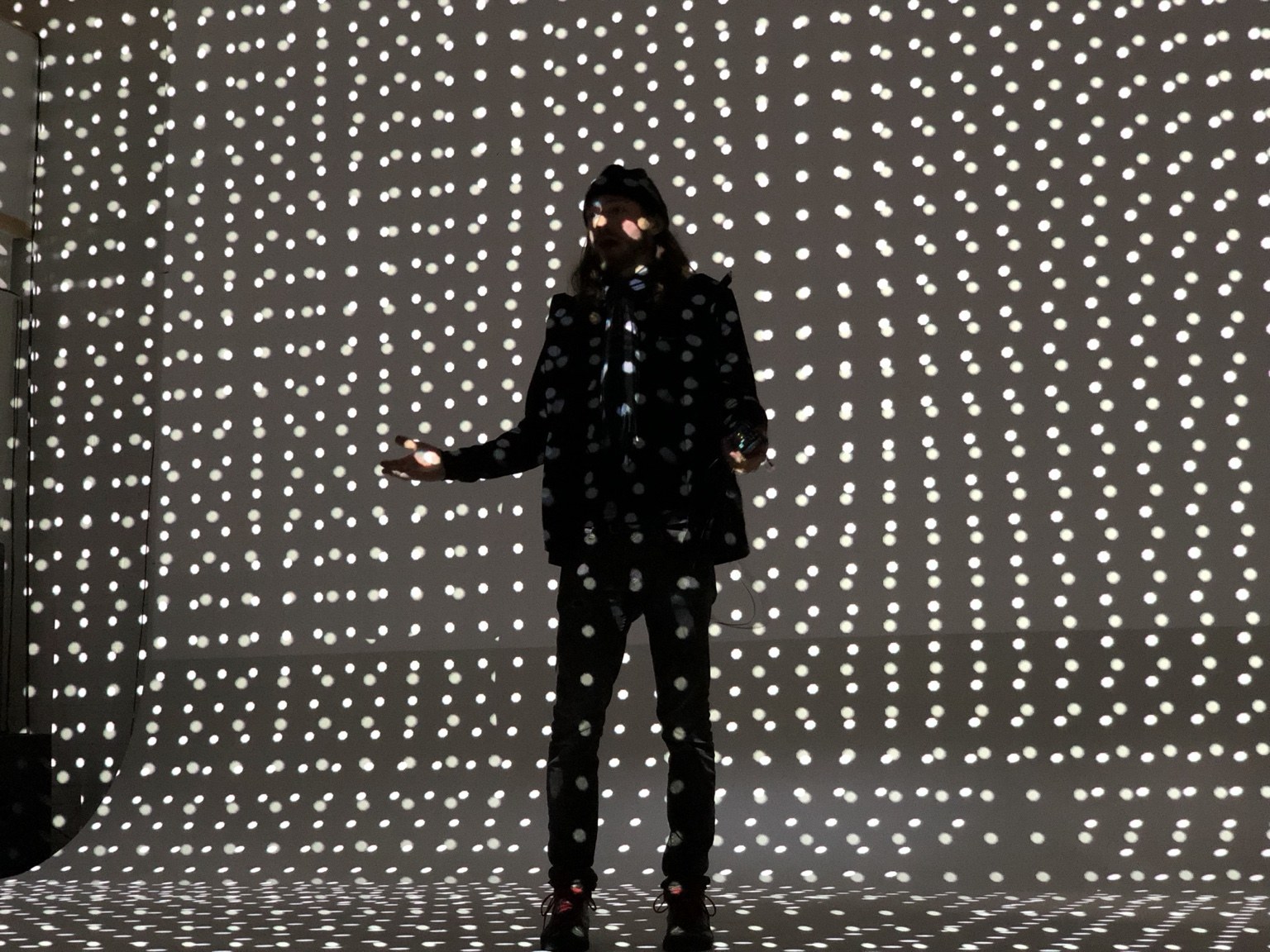 Tatton presenting with a microphone while a white dot pattern is projected across him, the wall, and the floor.